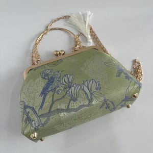 Peut inclure: Un sac à main vert clair orné d'un motif d'oiseau bleu et de fleurs, doté d'une armature en métal doré avec un fermoir à pression, d'une bandoulière en chaîne dorée amovible et d'un pompon blanc. La base comporte quatre petits pieds dorés.