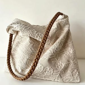 Puede incluir: Bolso tote color crema con intrincado bordado floral. El bolso presenta una correa trenzada de cuero marrón. La tela tiene una apariencia delicada y texturizada, y el diseño es una mezcla de estilos clásicos y bohemios.