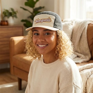 May include: A beige and gray baseball cap with the word "Venezuela" in a script font. The cap has a flat brim and a five-panel design. The cap is worn by a person.