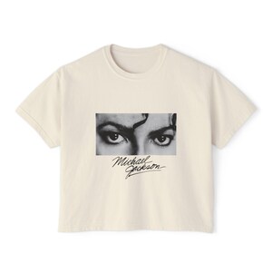 May include: A cream-coloured t-shirt with a black and white image of Michael Jackson's eyes. The singer's signature is printed below the image. The shirt has a crew neck and short sleeves.