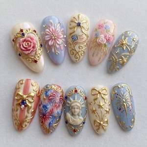 May include: A collection of ten intricately designed artificial nails. Each nail features a unique design, including floral patterns, bows, stars, and the American flag, all in a palette of gold, red, blue, and white. The nails are oval-shaped and appear to be professionally crafted.