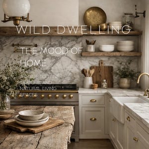 Könnte beinhalten: Ein gebundenes Buch mit dem Titel WILD DWELLING THE MOOD OF HOME A GUIDE TO TONE, TEXTURE, AND NATURAL LIVING SPACES Volume I. Das Cover zeigt eine Küche mit einem rustikalen Holztisch, Marmorarbeitsplatten, weißen Schränken und Messingbeschlägen.