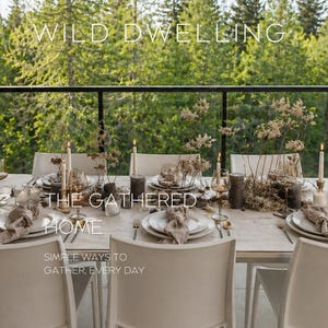 May include: A table setting for a gathering, with white plates, beige napkins, and lit candles. Silverware and glassware are arranged on the table. The text "WILD DWELLING" and "THE GATHERED HOME" are visible. A green forest forms the backdrop.