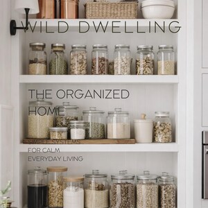 May include: A white built-in shelving unit featuring three levels of clear glass storage jars filled with various dry goods, copper canisters, a woven basket, and white bowls. The text WILD DWELLING THE ORGANIZED HOME SIMPLE SYSTEMS FOR CALM EVERYDAY LIVING Volume III is overlaid on the image.