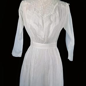 May include: A vintage white dress with long sleeves and a high, lace-trimmed neckline. The dress features vertical pleats on the bodice and skirt, and a defined waist. The dress is displayed against a black background.