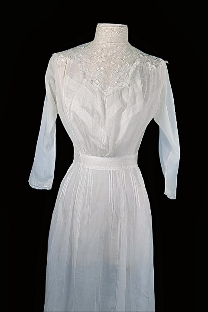 May include: A vintage white dress with long sleeves and a high, lace-trimmed neckline. The dress features vertical pleats on the bodice and skirt, and a defined waist. The dress is displayed against a black background.