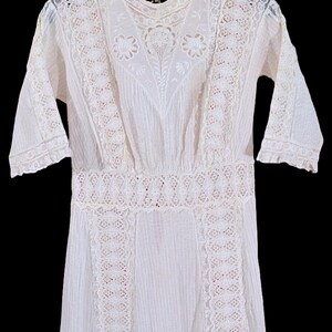 May include: A white vintage dress with intricate lace detailing. The dress features a high neckline, short sleeves, and a fitted waist. The lace is used on the bodice, sleeves, and hem, creating a delicate and elegant look. The dress is hanging on a black hanger.