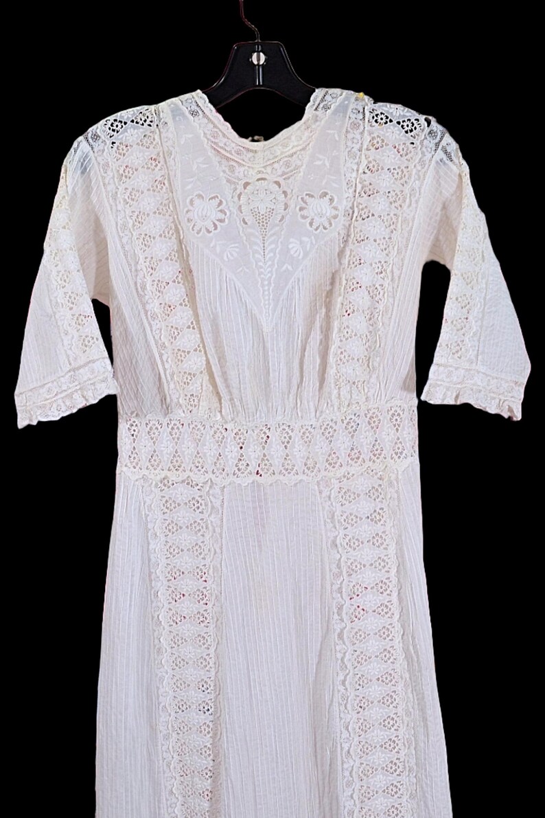May include: A white vintage dress with intricate lace detailing. The dress features a high neckline, short sleeves, and a fitted waist. The lace is used on the bodice, sleeves, and hem, creating a delicate and elegant look. The dress is hanging on a black hanger.