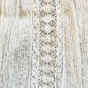 May include: Close-up of a white fabric with vertical ribbed texture and floral lace detailing. The lace is centered and features a repeating pattern of small flowers. The fabric appears to be cotton or a similar lightweight material.