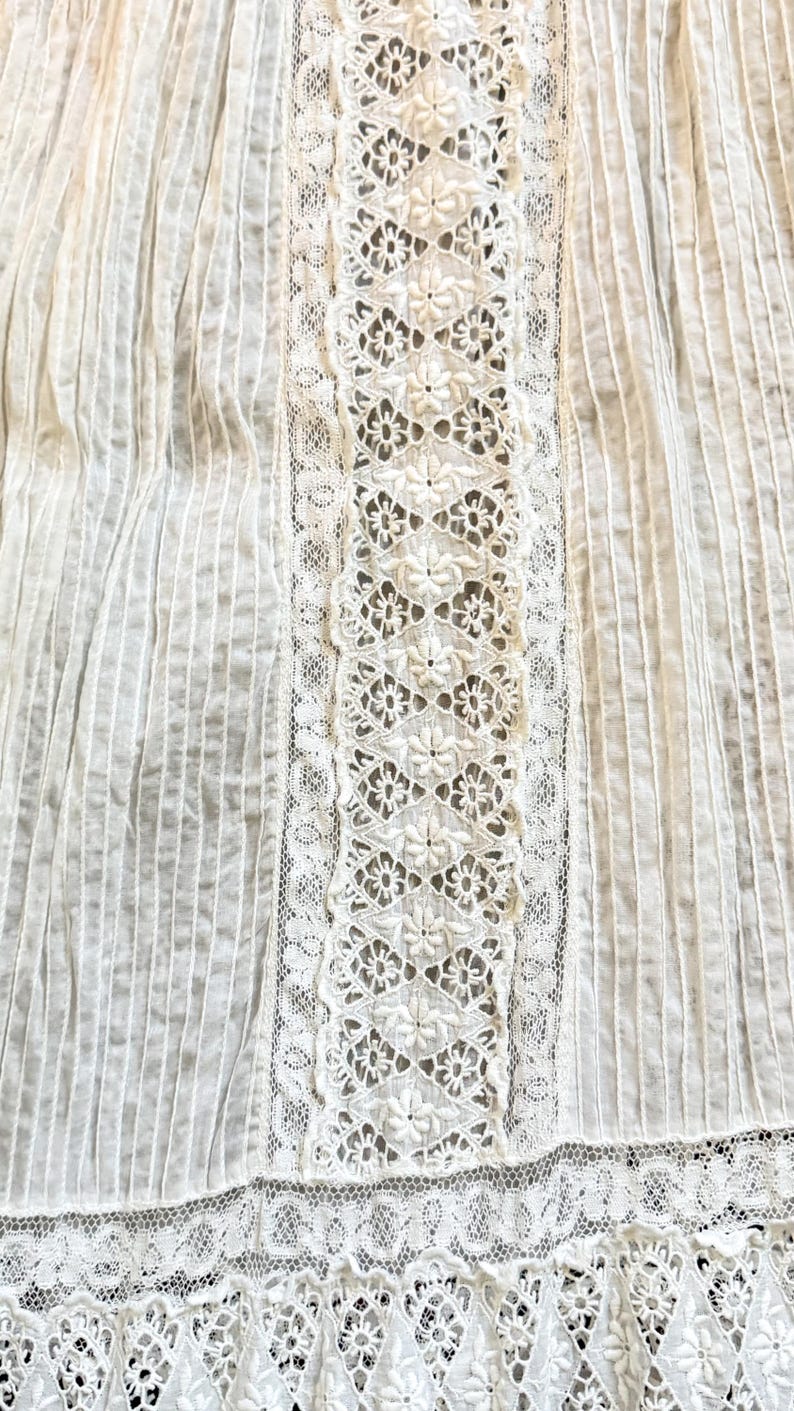 May include: Close-up of a white fabric with vertical ribbed texture and floral lace detailing. The lace is centered and features a repeating pattern of small flowers. The fabric appears to be cotton or a similar lightweight material.