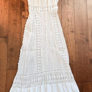 May include: A white, long-sleeved dress with intricate lace detailing. The dress features a high neckline, fitted bodice, and a tiered skirt. The fabric appears to be lightweight, with vertical stripes and lace inserts. The dress is laid out on a wooden surface.