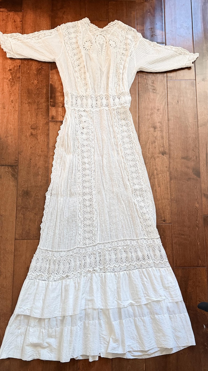 May include: A white, long-sleeved dress with intricate lace detailing. The dress features a high neckline, fitted bodice, and a tiered skirt. The fabric appears to be lightweight, with vertical stripes and lace inserts. The dress is laid out on a wooden surface.