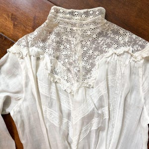May include: Close-up of a white vintage blouse with intricate lace detailing. The blouse features a high, lace-trimmed collar and delicate lace inserts on the bodice. The fabric appears to be lightweight and semi-sheer, with a classic, elegant design.