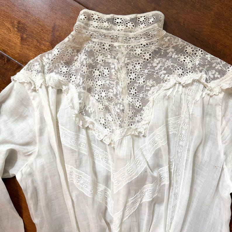 May include: Close-up of a white vintage blouse with intricate lace detailing. The blouse features a high, lace-trimmed collar and delicate lace inserts on the bodice. The fabric appears to be lightweight and semi-sheer, with a classic, elegant design.
