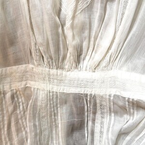 May include: Close-up of a white, vintage-style blouse. The fabric is sheer and features vertical pleats and decorative horizontal bands with intricate patterns. The sleeves are visible on the left side.