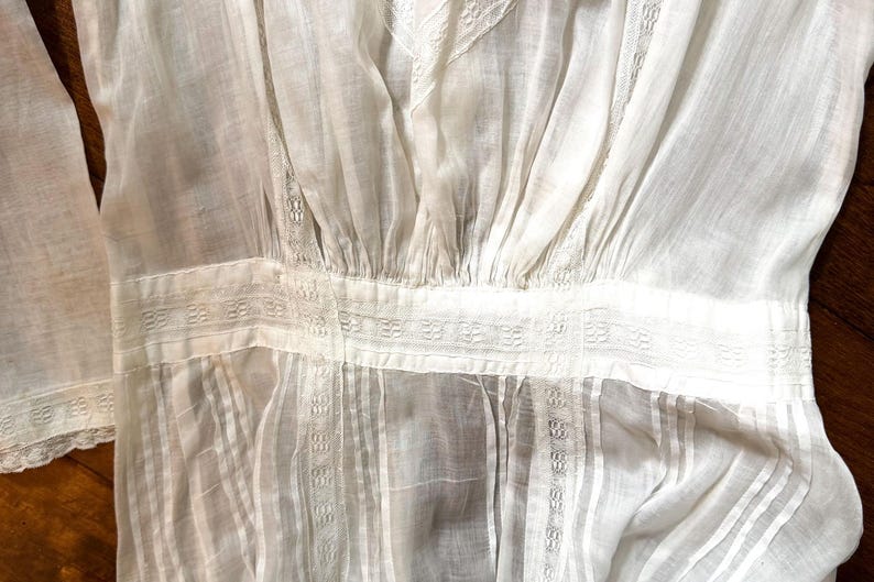 May include: Close-up of a white, vintage-style blouse. The fabric is sheer and features vertical pleats and decorative horizontal bands with intricate patterns. The sleeves are visible on the left side.