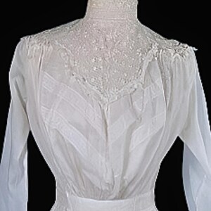 May include: A vintage white dress with long sleeves and a high, lace-trimmed collar. The dress features intricate detailing on the bodice and a fitted waist. The fabric appears to be lightweight and semi-sheer, with a flowing skirt.