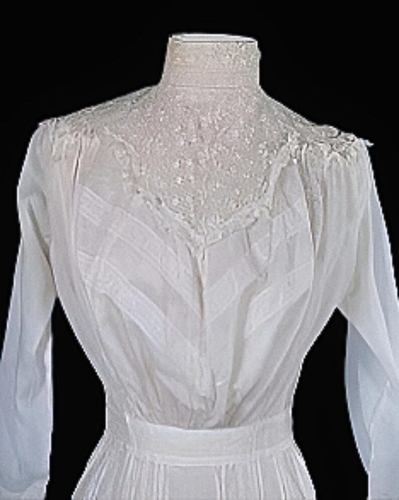 May include: A vintage white dress with long sleeves and a high, lace-trimmed collar. The dress features intricate detailing on the bodice and a fitted waist. The fabric appears to be lightweight and semi-sheer, with a flowing skirt.
