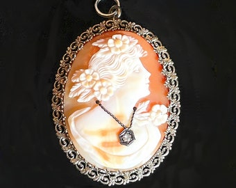 May include: Oval cameo pendant featuring a woman's profile carved in cream and peach tones, adorned with floral accents. The pendant is set in a decorative silver-toned frame and includes a small diamond pendant. A classic piece of jewelry.