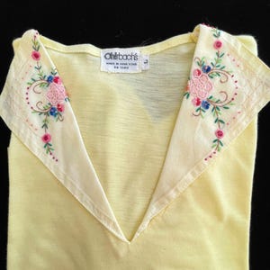 May include: A pale yellow vintage top with a V-neck and a decorative collar. The collar is off-white and features embroidered pink, blue, and green floral designs. The tag reads "Ohrbach's Made in Hong Kong".