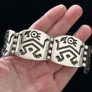 May include: Silver bracelet featuring a repeating design of stylized figures. The bracelet is composed of rectangular links with rounded edges, connected by small, round beads. The figures are black against a silver background, creating a striking contrast.