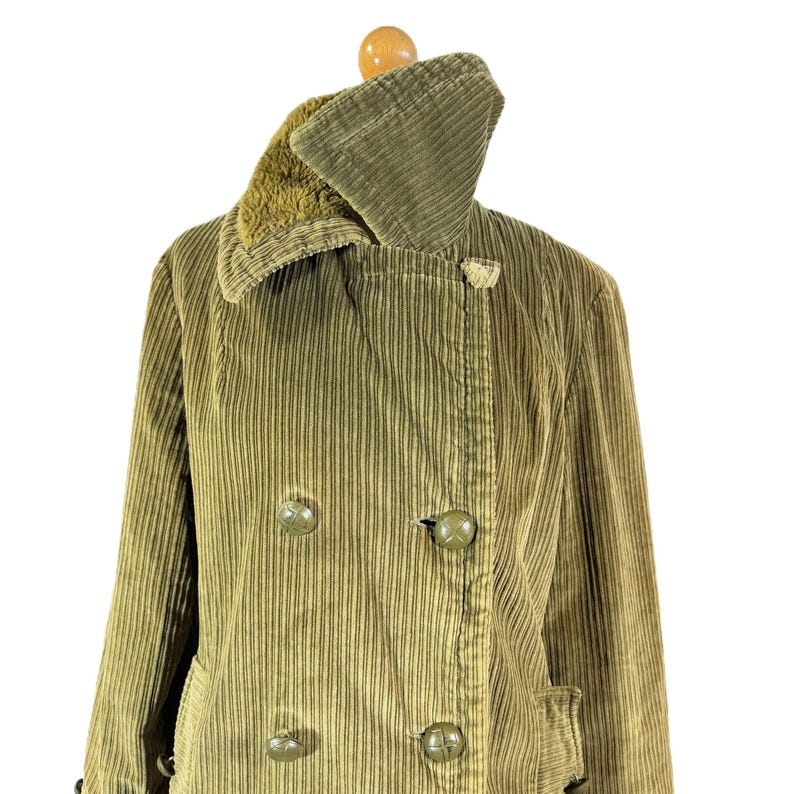 May include: Olive green corduroy coat with a double-breasted design. The coat features a wide collar with a faux fur lining and large, round buttons. The coat is displayed on a wooden hanger.