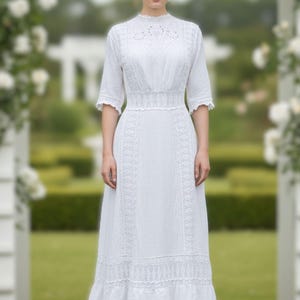 May include: A white, floor-length dress with a high neckline and short sleeves. The dress features intricate lace detailing on the bodice, sleeves, and hem. The dress is made of a lightweight fabric and has a fitted waist. The woman is standing in a garden setting.