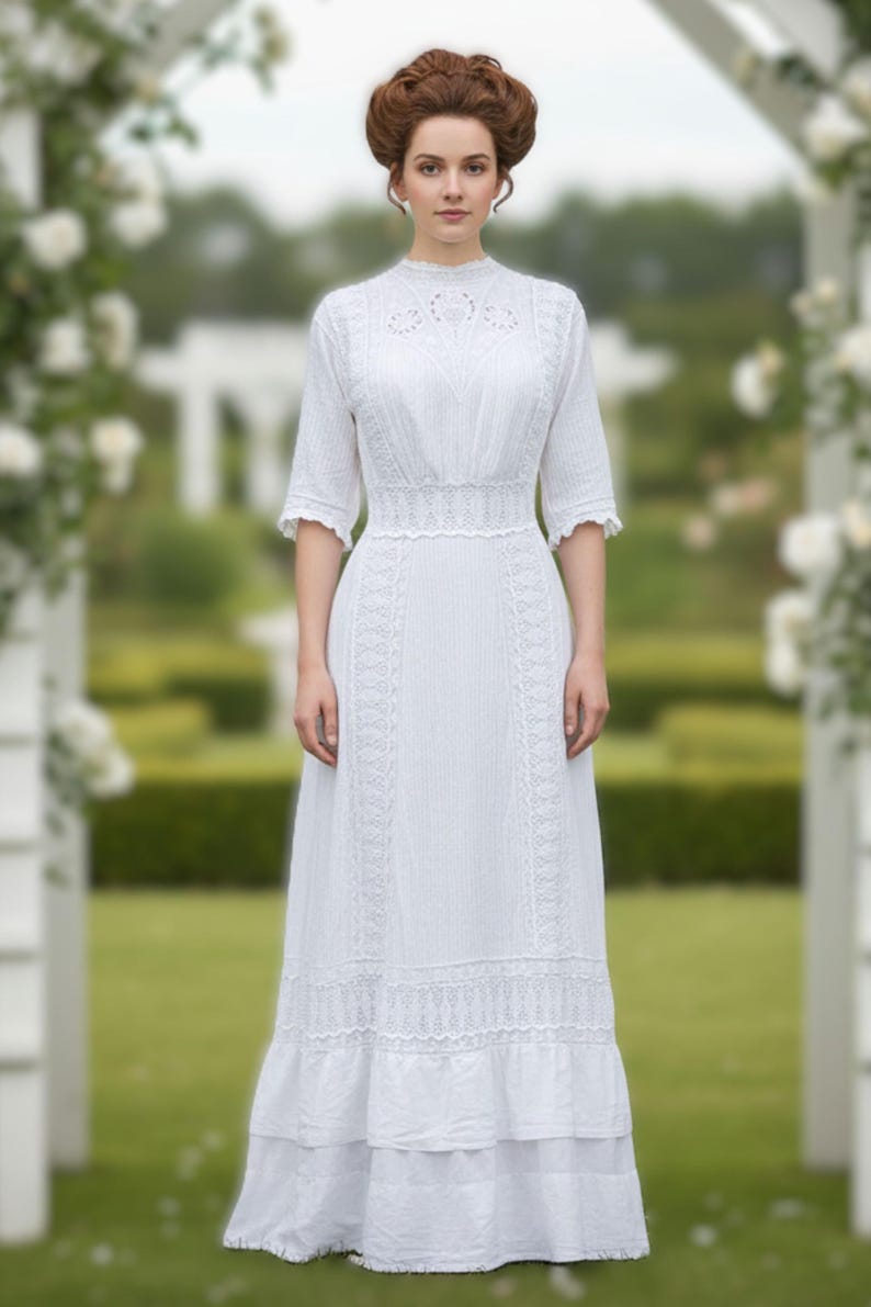 May include: A white, floor-length dress with a high neckline and short sleeves. The dress features intricate lace detailing on the bodice, sleeves, and hem. The dress is made of a lightweight fabric and has a fitted waist. The woman is standing in a garden setting.