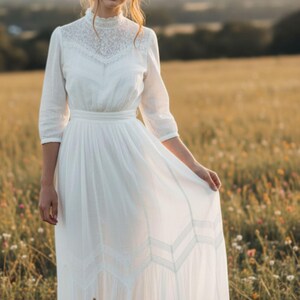 May include: A white dress with a high neckline and lace detailing. The dress has three-quarter sleeves and a flowing skirt. The person is standing in a field of wildflowers. The dress appears to be made of a lightweight fabric.