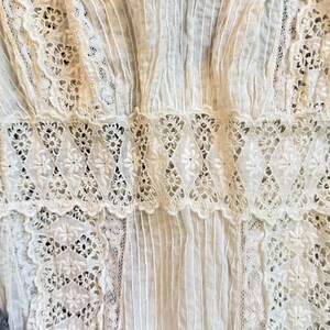 May include: Close-up of a white garment featuring intricate lace and pleating details. The design includes floral lace patterns and vertical pleats, creating a textured and delicate appearance. The garment appears to be made of a soft, lightweight fabric.