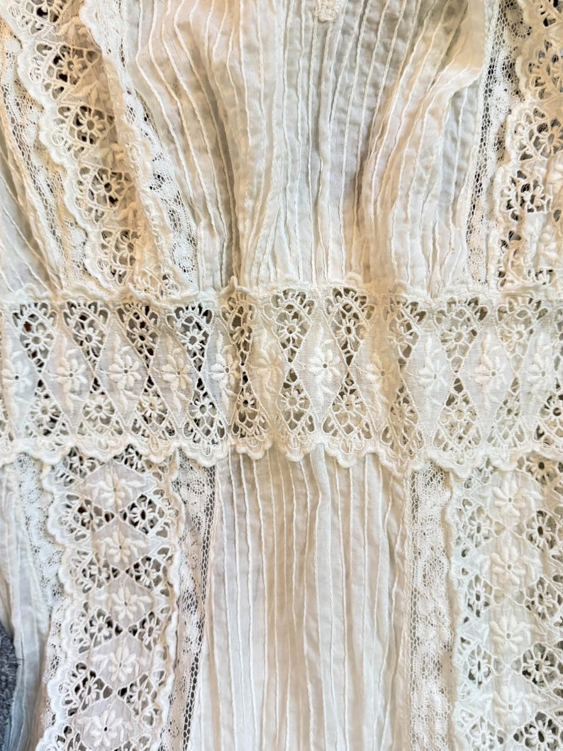 May include: Close-up of a white garment featuring intricate lace and pleating details. The design includes floral lace patterns and vertical pleats, creating a textured and delicate appearance. The garment appears to be made of a soft, lightweight fabric.