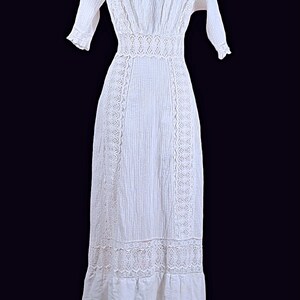 May include: A white, floor-length dress with intricate lace detailing. The dress features a high neckline, short sleeves, and a fitted waist. Vertical textured stripes run down the length of the dress, with lace accents along the sides and hem.