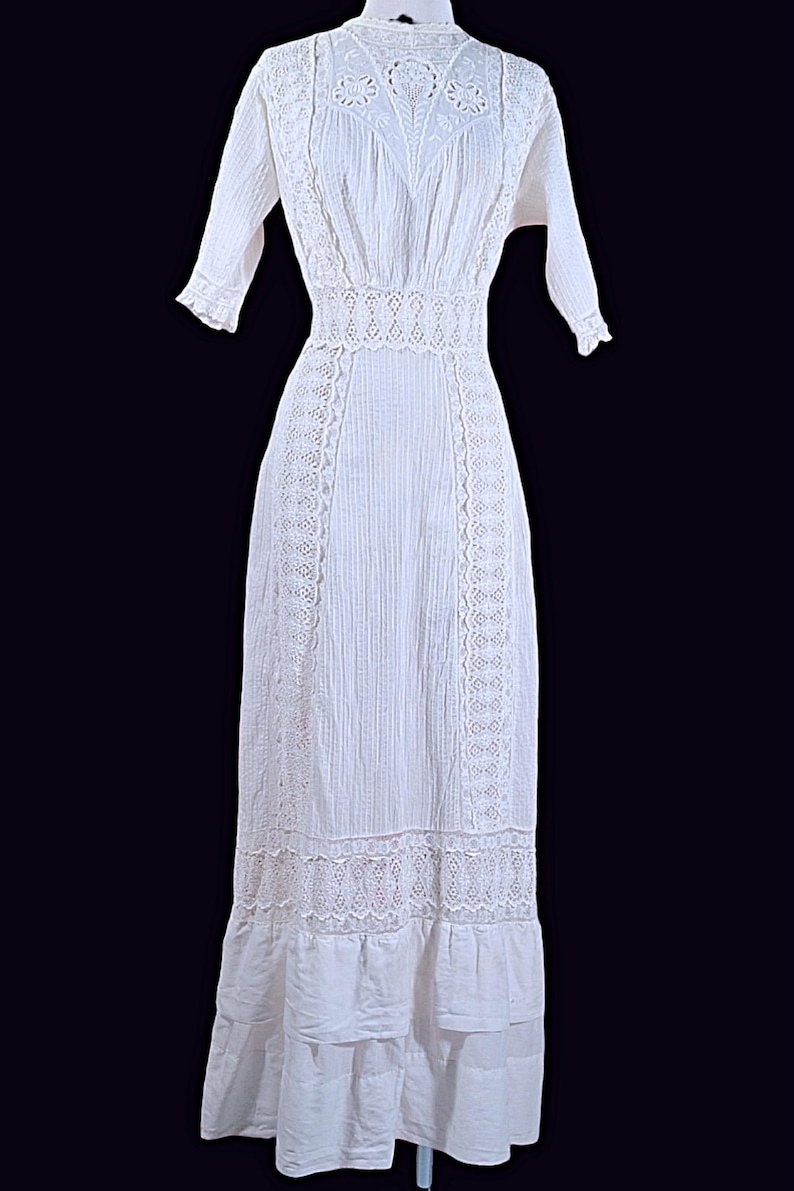 May include: A white, floor-length dress with intricate lace detailing. The dress features a high neckline, short sleeves, and a fitted waist. Vertical textured stripes run down the length of the dress, with lace accents along the sides and hem.