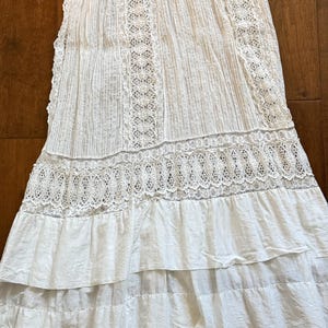 May include: White cotton skirt with intricate lace detailing. The skirt features vertical pleats and horizontal lace bands, with a tiered, ruffled hemline. The fabric appears lightweight and suitable for warm weather.