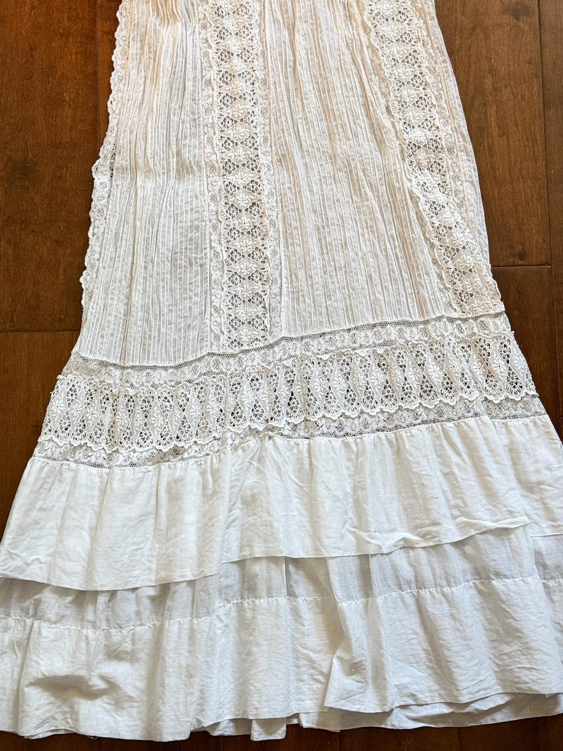 May include: White cotton skirt with intricate lace detailing. The skirt features vertical pleats and horizontal lace bands, with a tiered, ruffled hemline. The fabric appears lightweight and suitable for warm weather.
