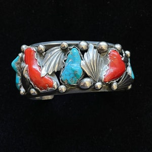 May include: A silver bracelet with turquoise and coral stones set in a decorative design. The bracelet is made of sterling silver and features a variety of shapes and sizes of stones.