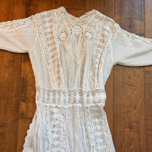 May include: A white vintage dress with intricate lace detailing. The dress features a high neckline, long sleeves, and a fitted waist. The bodice and sleeves have delicate lace inserts, and the skirt is pleated.