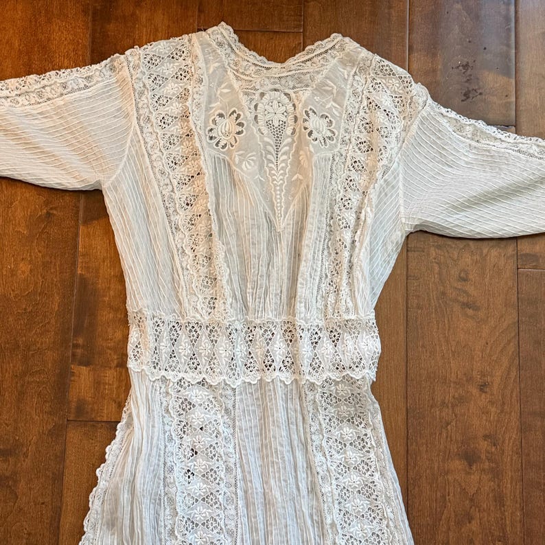 May include: A white vintage dress with intricate lace detailing. The dress features a high neckline, long sleeves, and a fitted waist. The bodice and sleeves have delicate lace inserts, and the skirt is pleated.