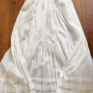 May include: A white, vintage-style dress with a flowing silhouette. The dress features intricate lace detailing and horizontal bands near the hem. The fabric appears lightweight and airy, suggesting a summer garment.