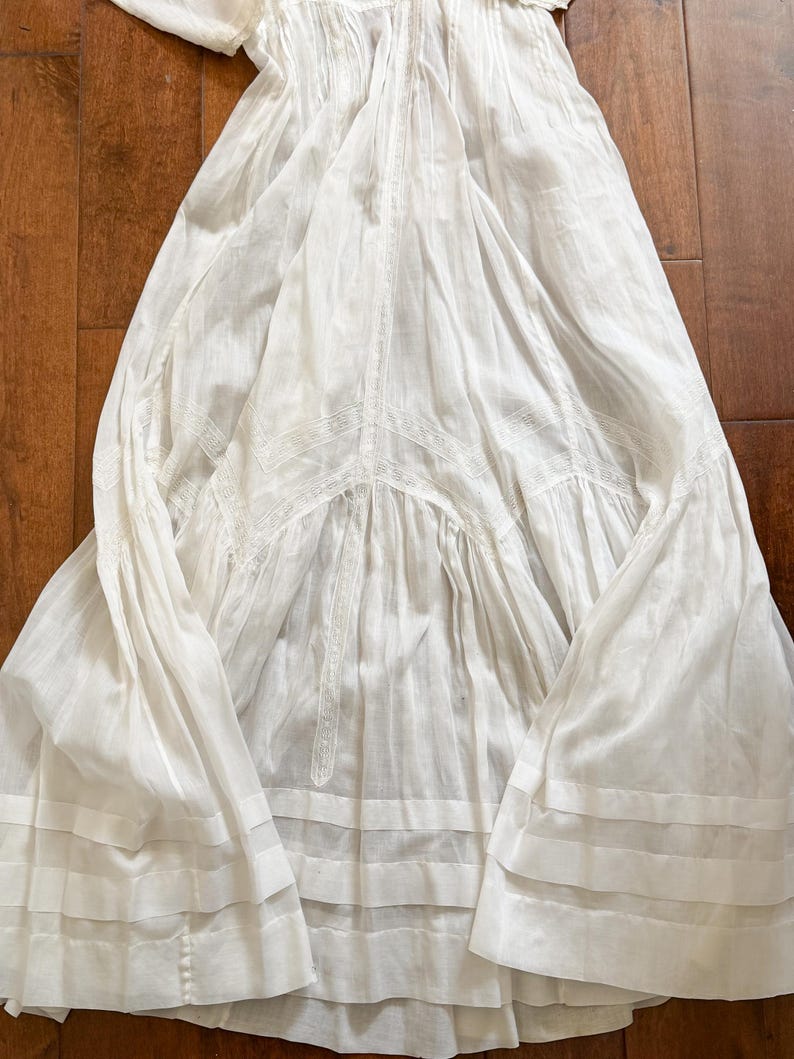 May include: A white, vintage-style dress with a flowing silhouette. The dress features intricate lace detailing and horizontal bands near the hem. The fabric appears lightweight and airy, suggesting a summer garment.