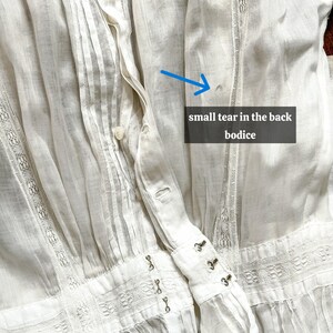 May include: Close-up of a white vintage blouse with intricate details. The blouse features pleats, lace trim, and hook-and-eye closures. The text in the image reads: "small tear in the back bodice."
