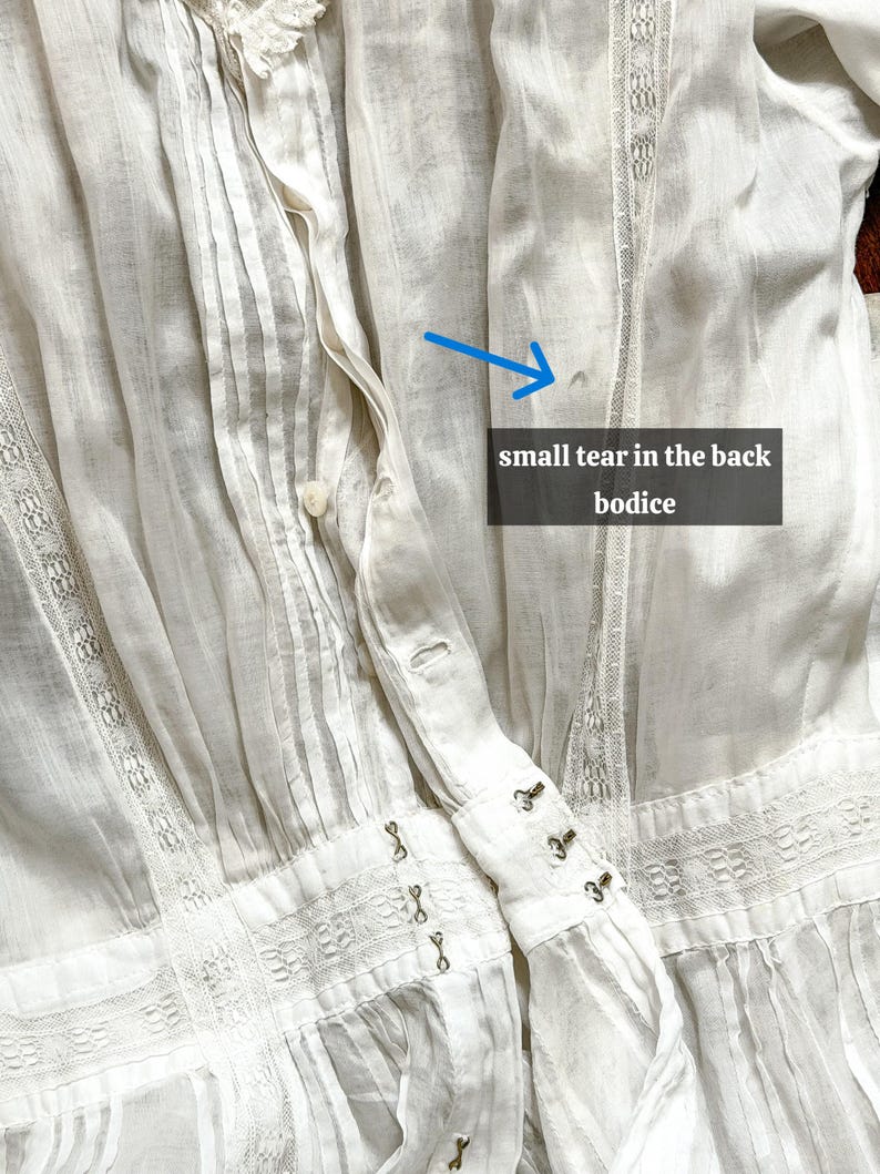 May include: Close-up of a white vintage blouse with intricate details. The blouse features pleats, lace trim, and hook-and-eye closures. The text in the image reads: "small tear in the back bodice."