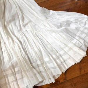 May include: A flowing white skirt with multiple layers and a tiered design. The skirt features delicate embroidery and horizontal bands near the hem. The fabric appears lightweight and airy, suggesting a summer or bohemian style.