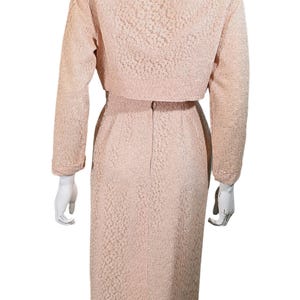 以下が含まれることがあります： マネキンに展示された、明るいピンク色のツーピーススーツ。長袖のクロップドジャケットと膝丈のペンシルスカートが含まれています。生地は、テクスチャ加工された花柄です。