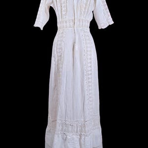 May include: A white vintage dress with a high neckline and short sleeves. The dress features intricate lace detailing along the bodice, sleeves, and hemline. The dress has a fitted waist and a flowing skirt, creating an elegant silhouette.