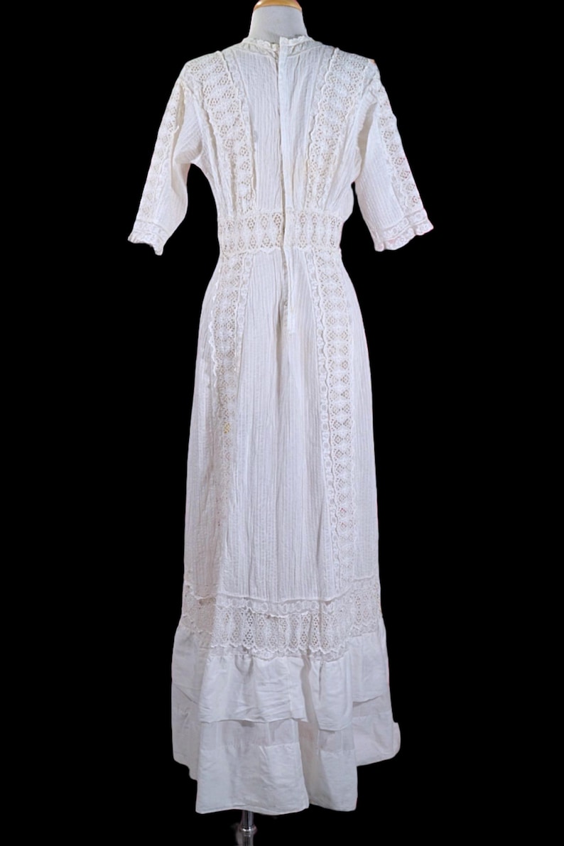 May include: A white vintage dress with a high neckline and short sleeves. The dress features intricate lace detailing along the bodice, sleeves, and hemline. The dress has a fitted waist and a flowing skirt, creating an elegant silhouette.