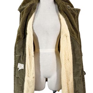 May include: Olive green corduroy jacket with a cream-colored, plush interior lining. The jacket is open, revealing the soft, warm lining and the texture of the corduroy fabric. The jacket is displayed on a mannequin.