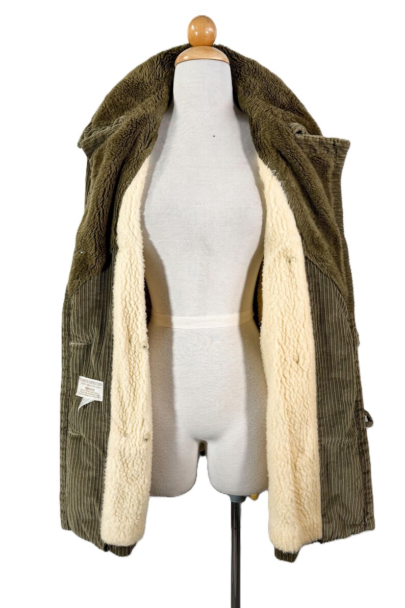 May include: Olive green corduroy jacket with a cream-colored, plush interior lining. The jacket is open, revealing the soft, warm lining and the texture of the corduroy fabric. The jacket is displayed on a mannequin.