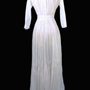 May include: A long, white dress with long sleeves and a high, lace-trimmed collar. The dress features a fitted waist and a flowing skirt, with vertical pleats and decorative panels near the hem. The dress is displayed against a black background.