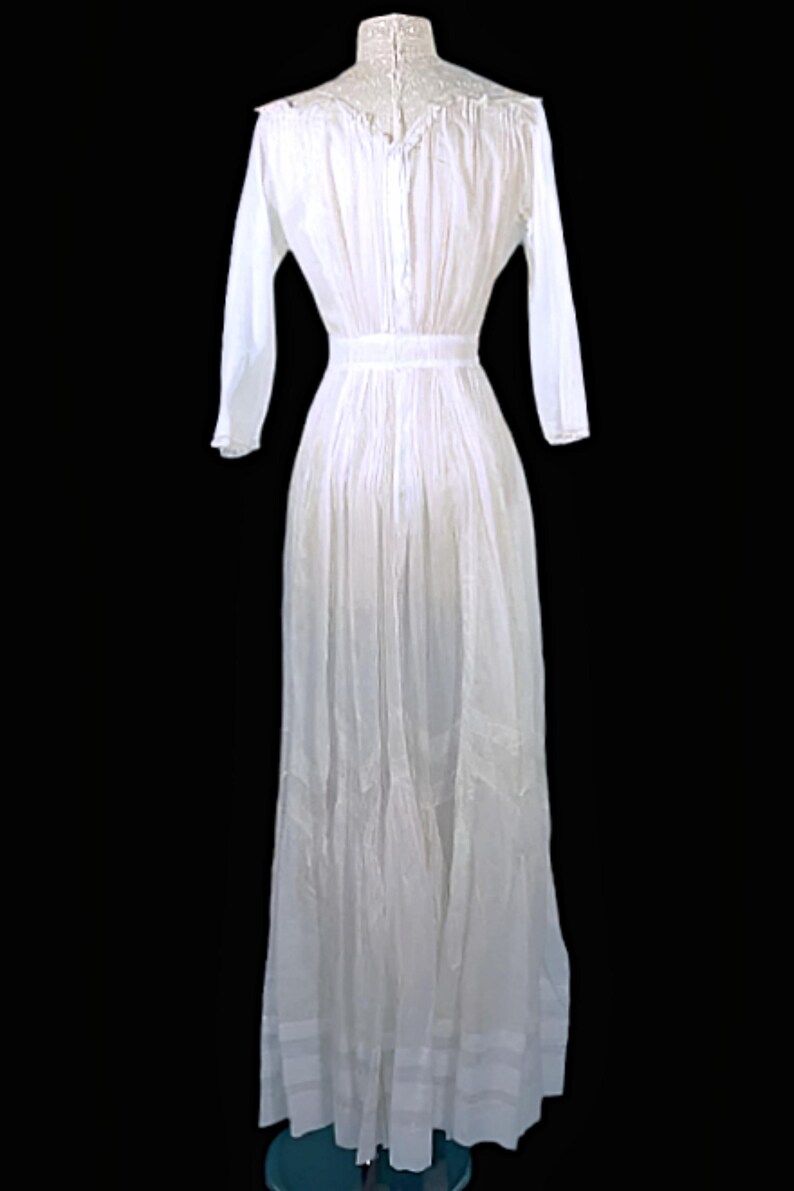 May include: A long, white dress with long sleeves and a high, lace-trimmed collar. The dress features a fitted waist and a flowing skirt, with vertical pleats and decorative panels near the hem. The dress is displayed against a black background.